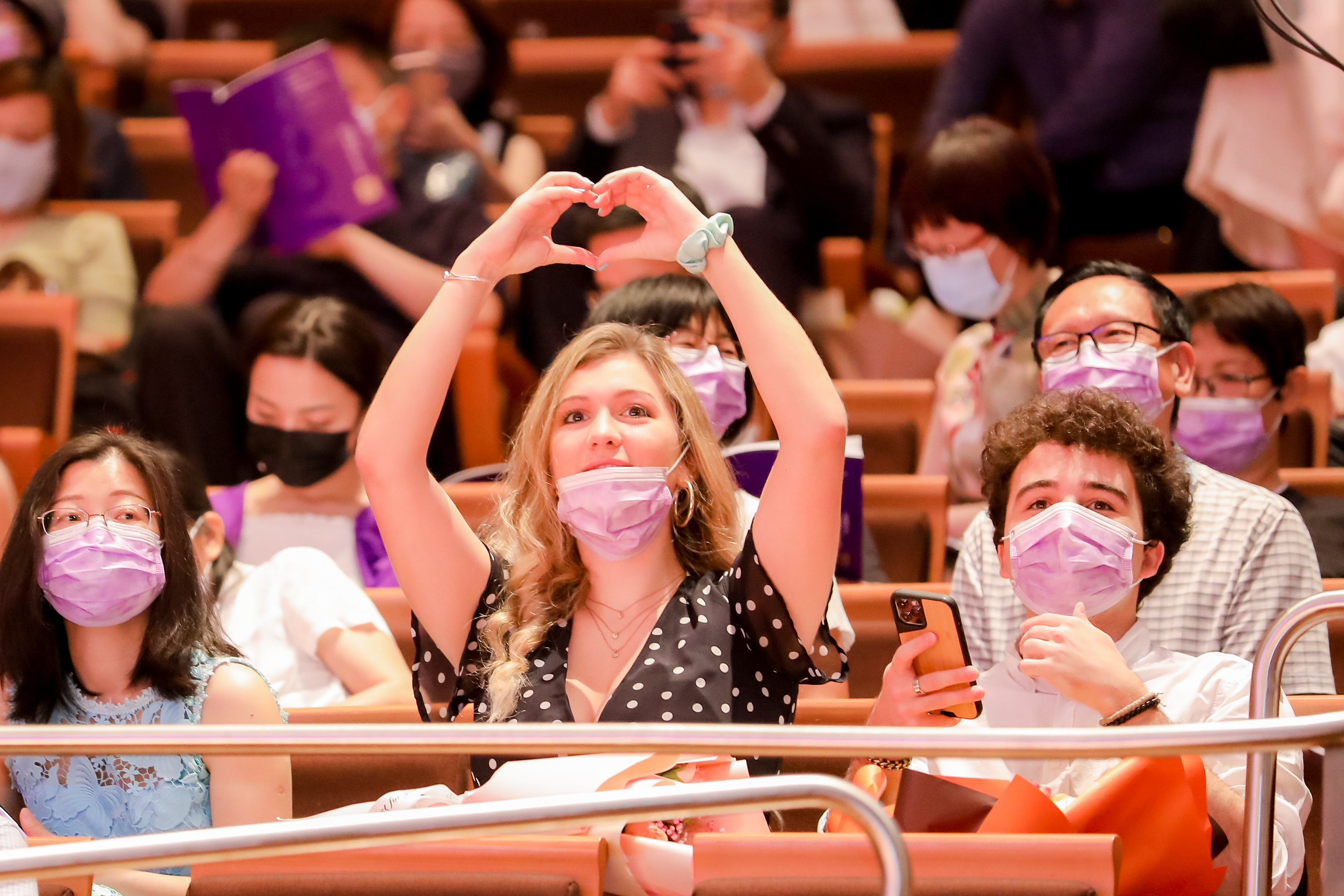 Young woman in audience raises her hands in a heart gesture toward graduates 