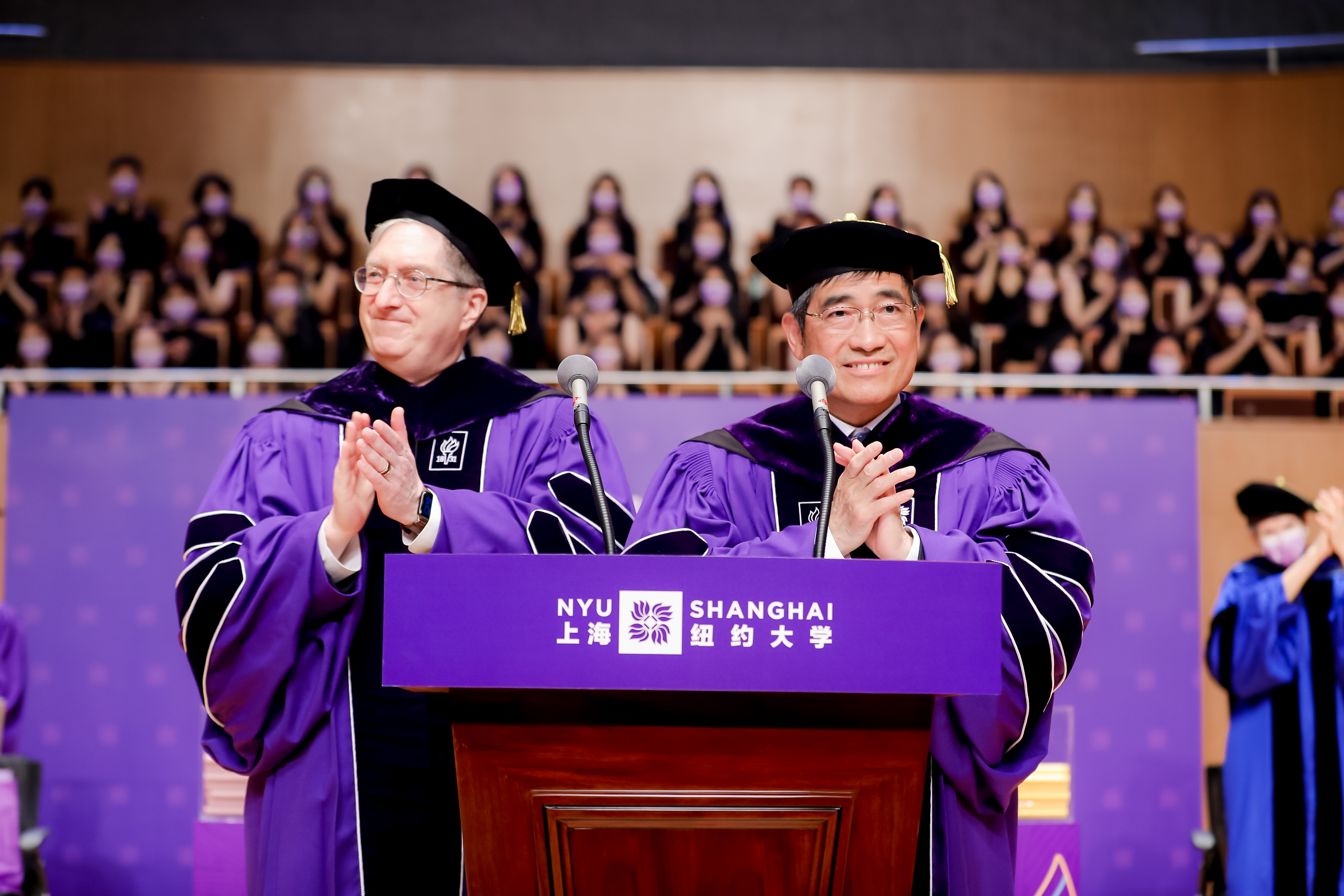 University leaders in purple robes applaud graduates at podium