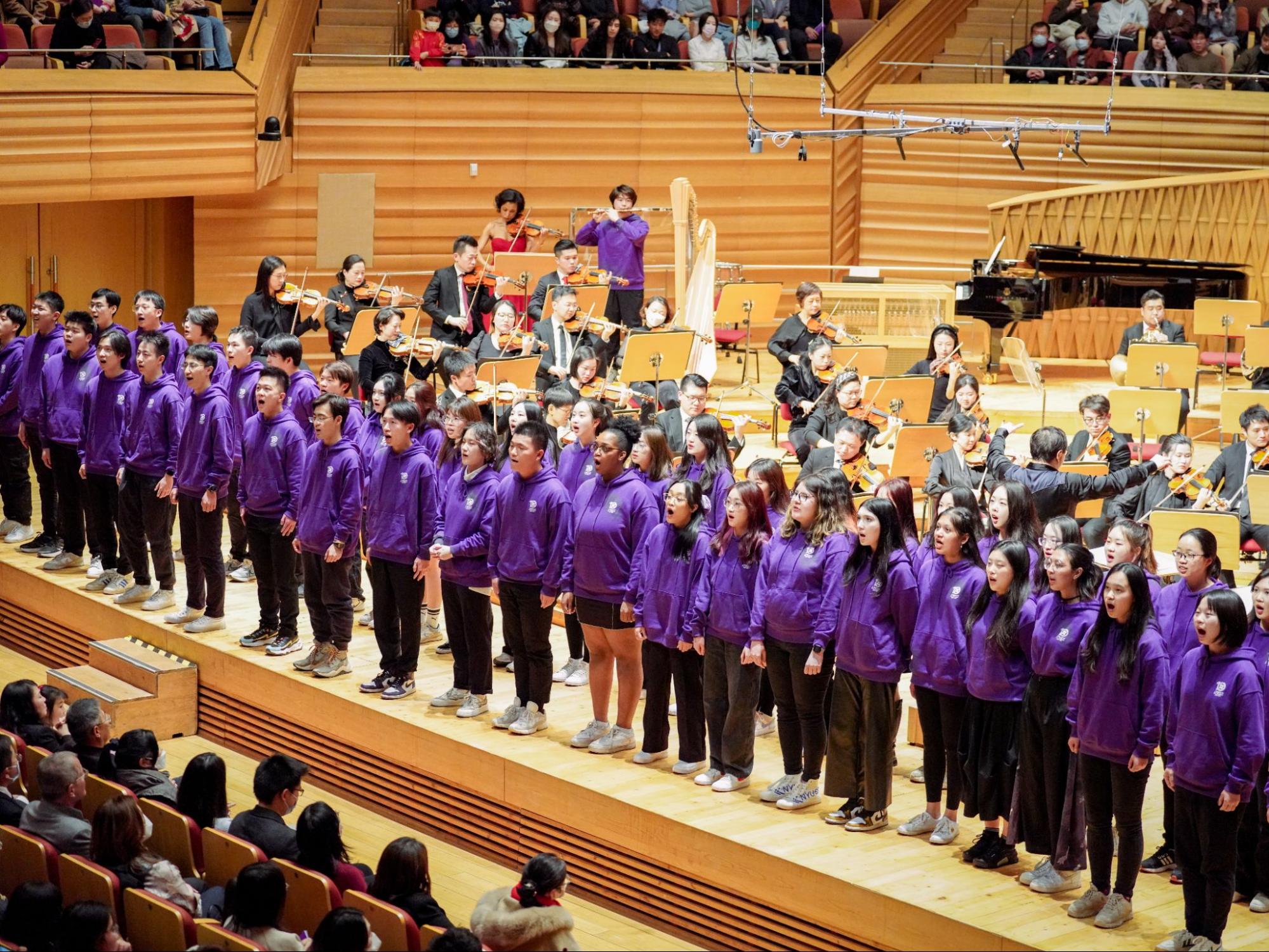 NYU Shanghai Chamber Singers stand on stage, singing in purple hoodies with the symphony behind them 
