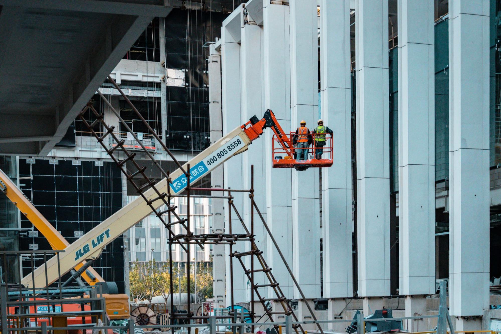 two construction workers stand on a crane overlooking the construction site.