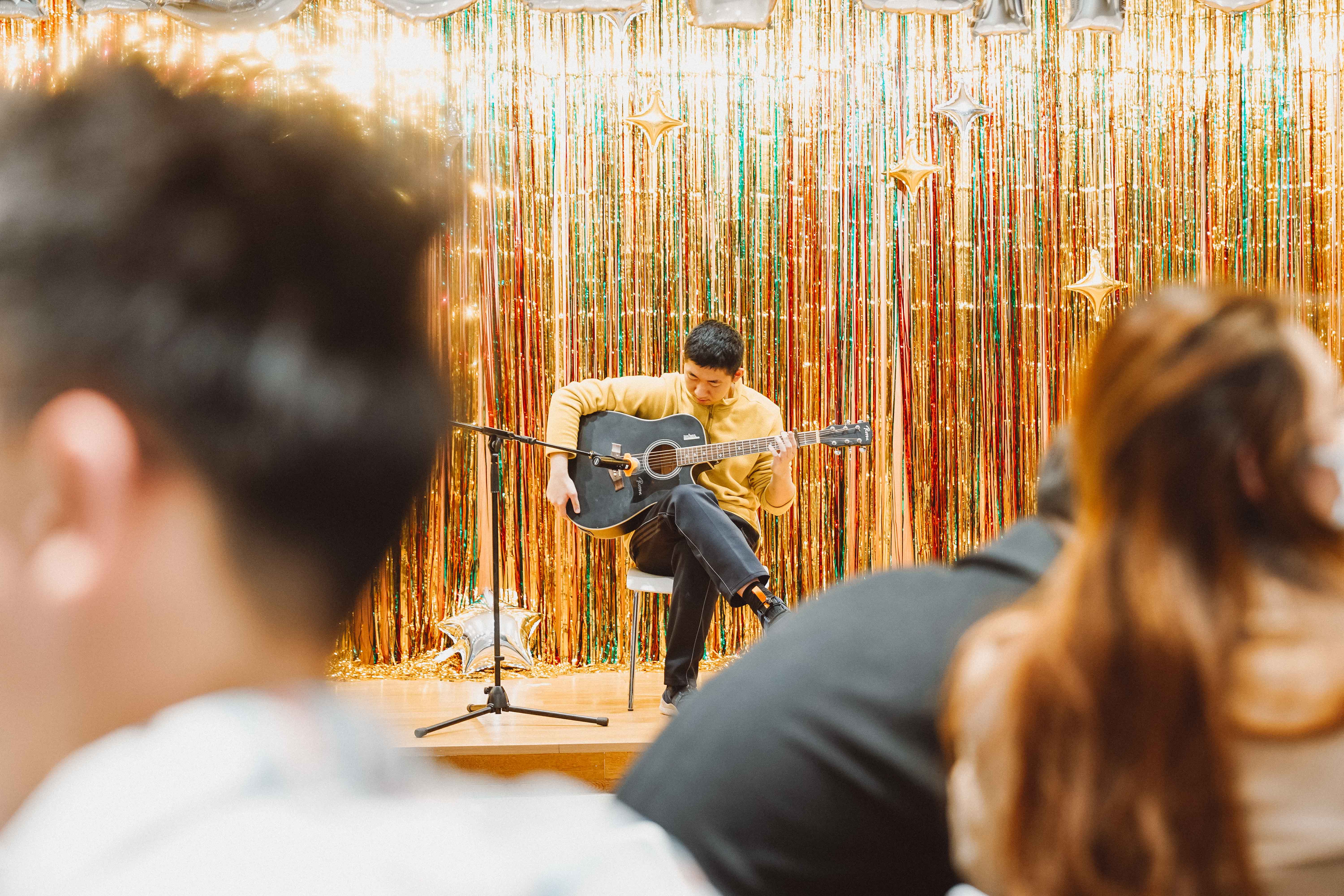 Daniel, a sophomore plays guitar on a stage with a sparkly backdrop. 