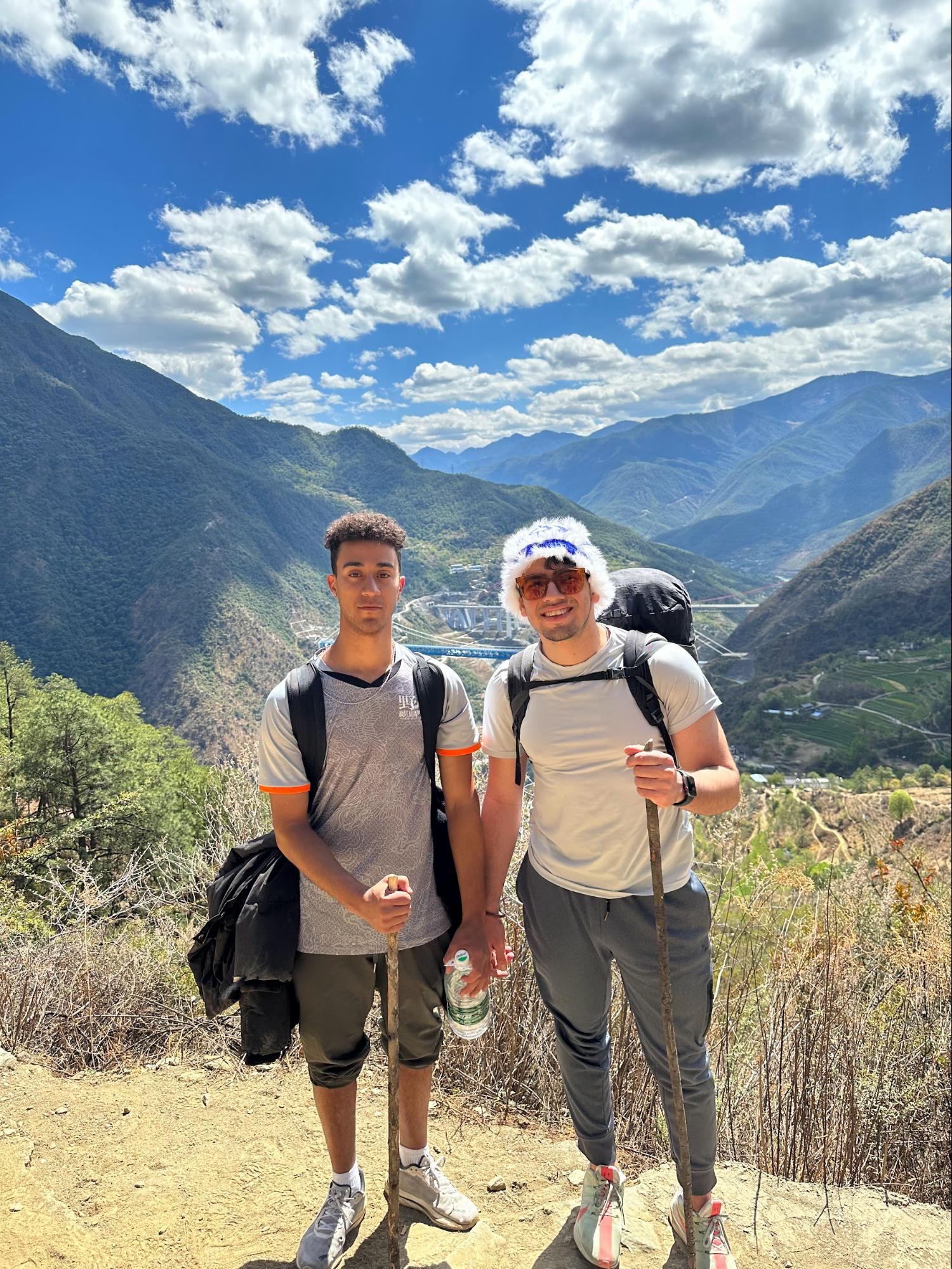 Two men pose with hiking sticks in front of a background of mountains and a cloudy sky