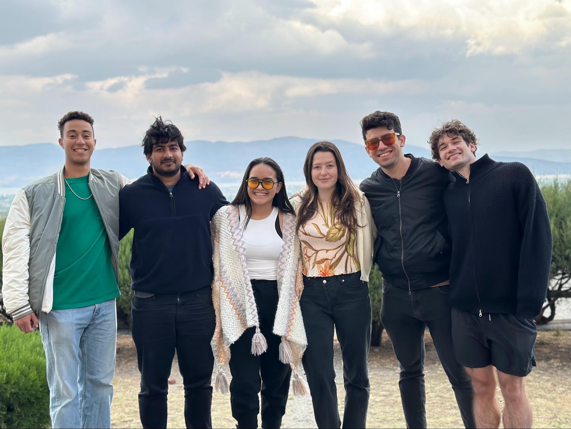 Six friends pose for a picture in front of a landscape of mountains and a cloudy sky