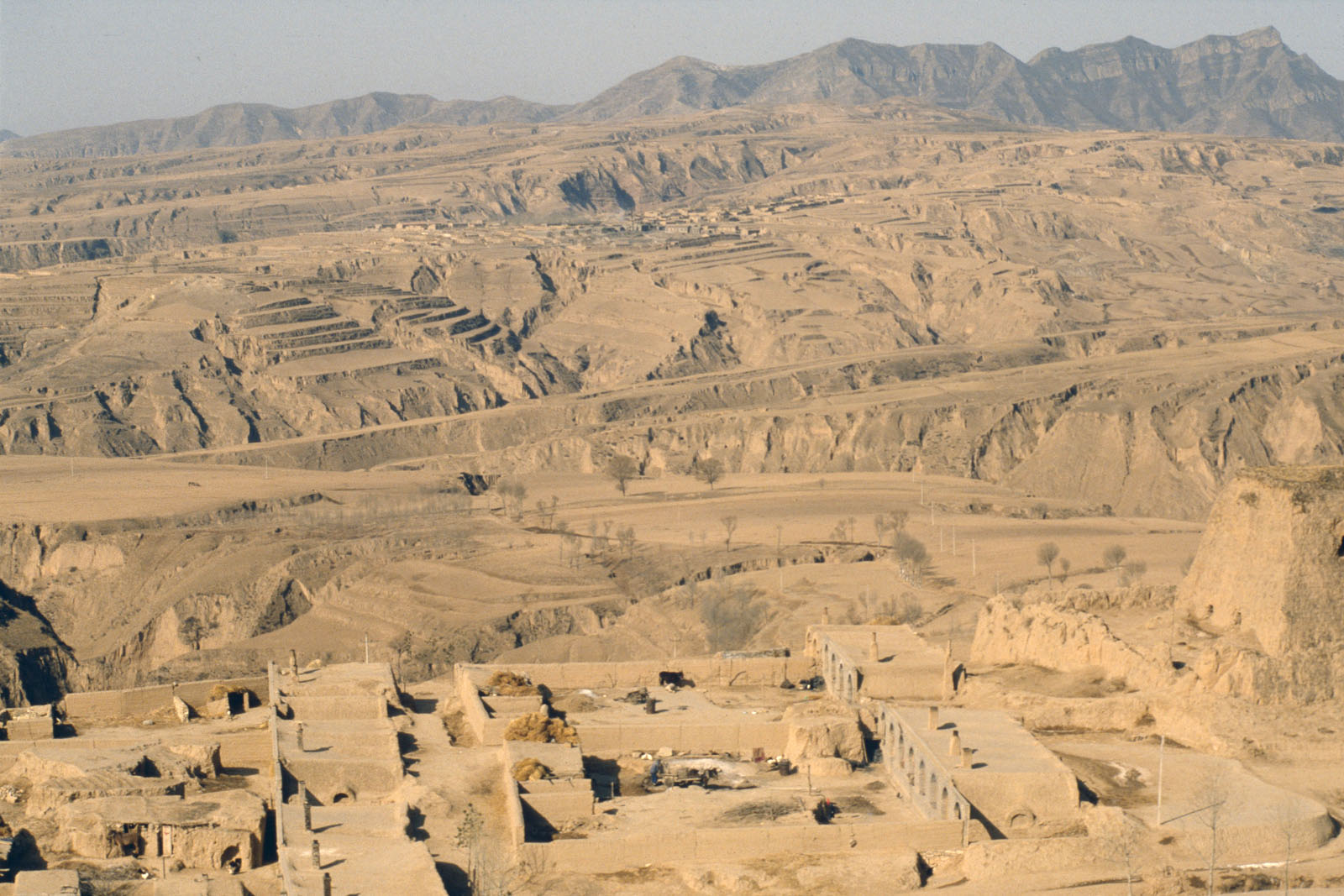 Brown and arid landscape of bare sandy hills and gullies with mountains in distance