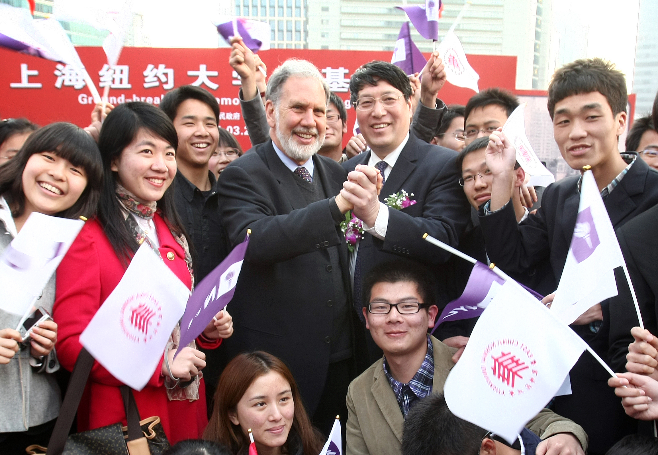 Sexton and Yu clasp hands surrounded by students waving NYU and ECNU flags