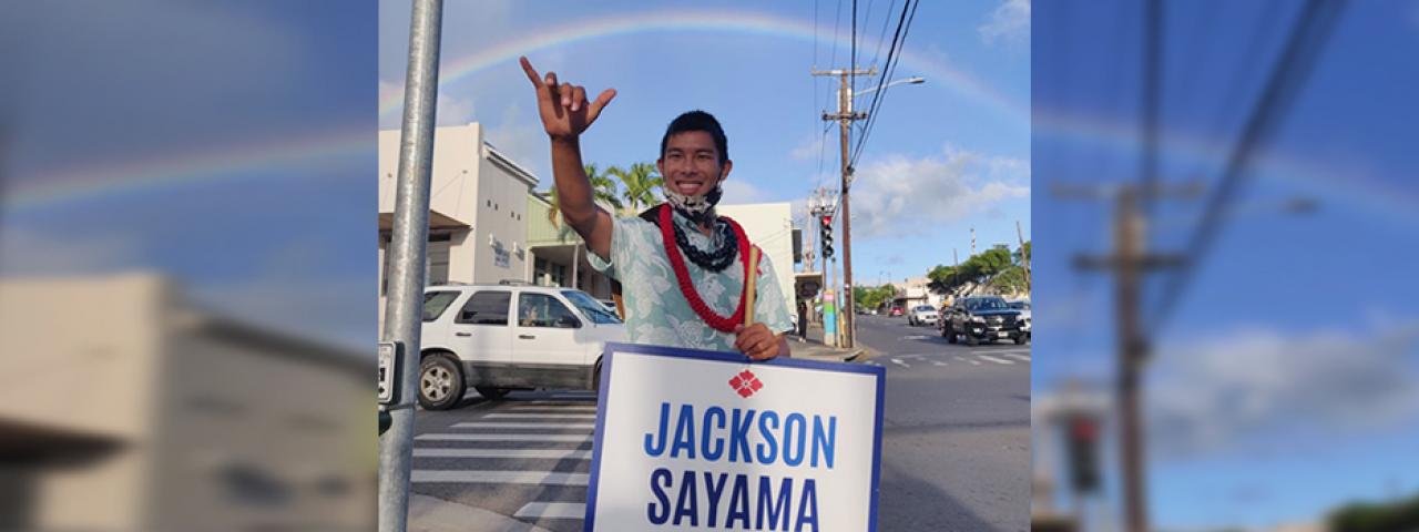 Sayama waves a sign under a rainbow