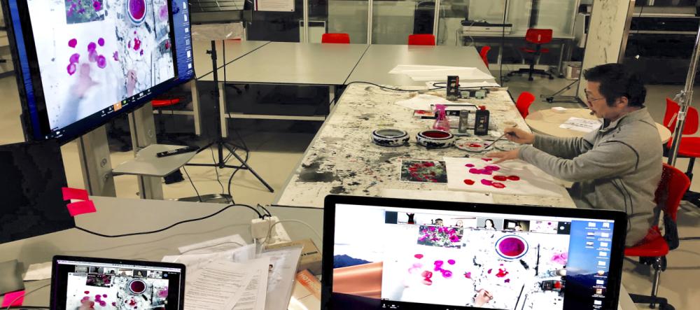 Jian-jun Zhang paints in classroom while multiple video feeds show both his hands and face on screen simultaneously